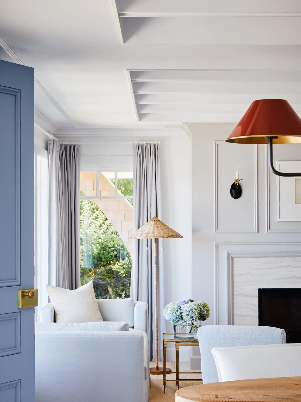 Bright living room with light blue armchairs, a blue door, wall molding, a fireplace, a red wall sconce, a floor lamp with a woven shade, and a vase of flowers on a side table.