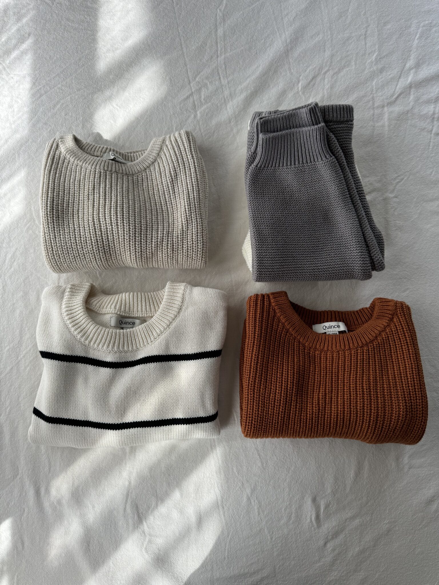 Four folded sweaters in different colors—beige, gray, white with black stripes, and burnt orange—are arranged on a white surface with sunlight casting soft shadows.