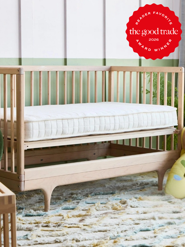A light wood crib with a mattress sits on a patterned rug in a nursery. A red award badge in the top right corner reads "The Good Trade Award Winner 2026".