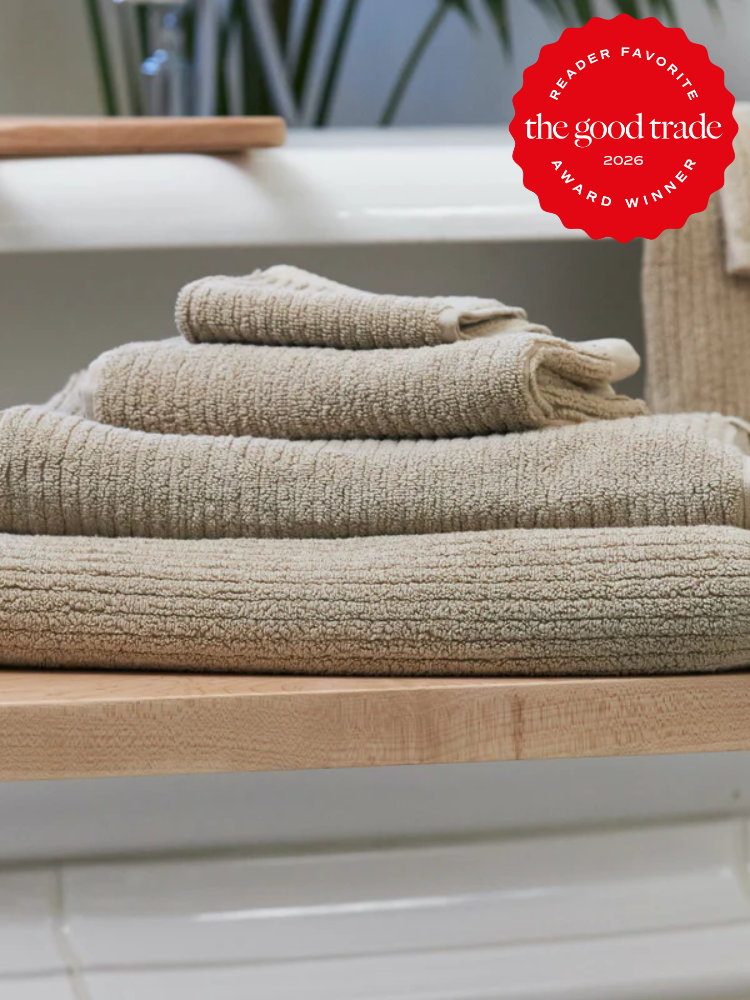 A stack of beige organic towels is neatly folded on a wooden shelf, with a red "The Good Trade Reader Favorite Award Winner 2026" badge in the upper right corner.