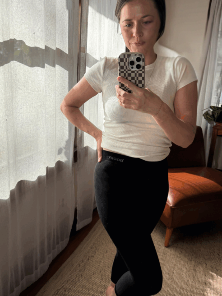 A person wearing a white t-shirt and black leggings takes a mirror selfie in a sunlit room with sheer curtains and a brown chair.