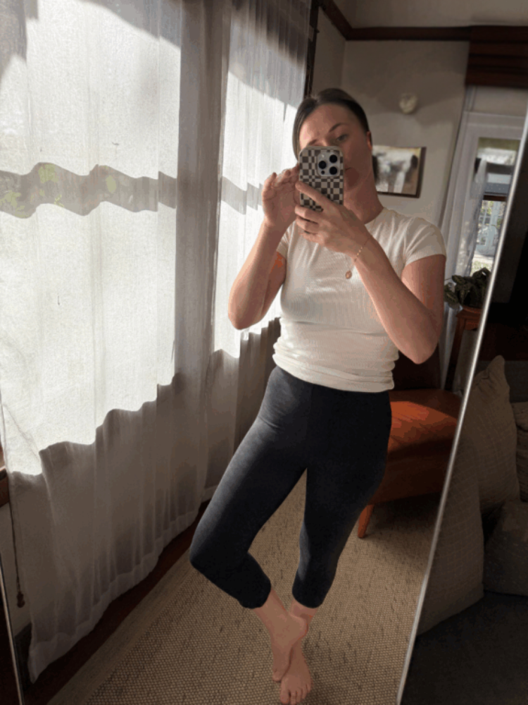 A person stands barefoot indoors, taking a mirror selfie. They are wearing a white top and black leggings in a sunlit room with sheer curtains and an orange chair.