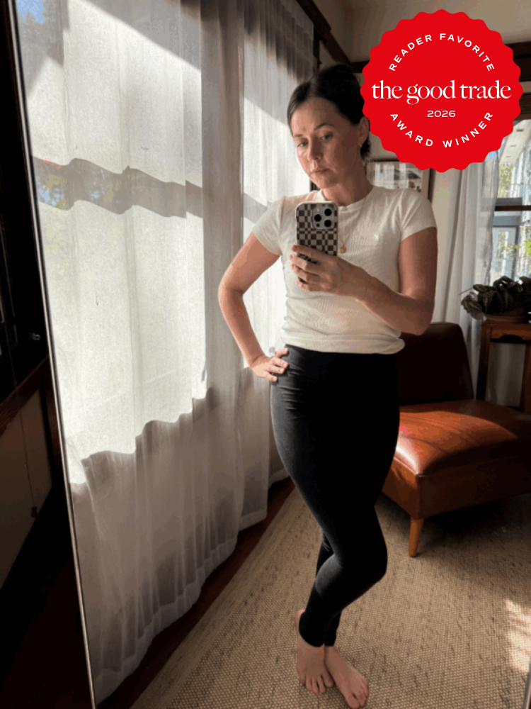 Woman stands barefoot indoors, taking a mirror selfie in a white top and black leggings; a "The Good Trade 2026 Award Winner" badge is displayed in the upper right corner.