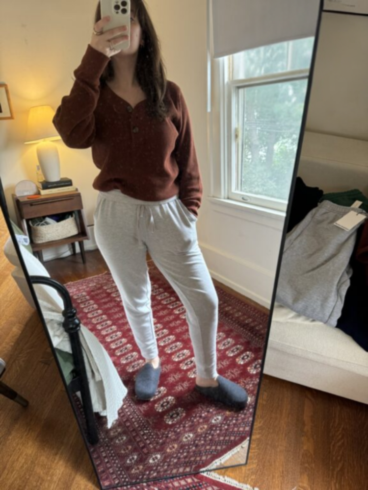 A person stands in front of a tall mirror wearing a brown sweater, light gray sweatpants, and dark slippers in a bedroom with wooden floors and a red patterned rug.