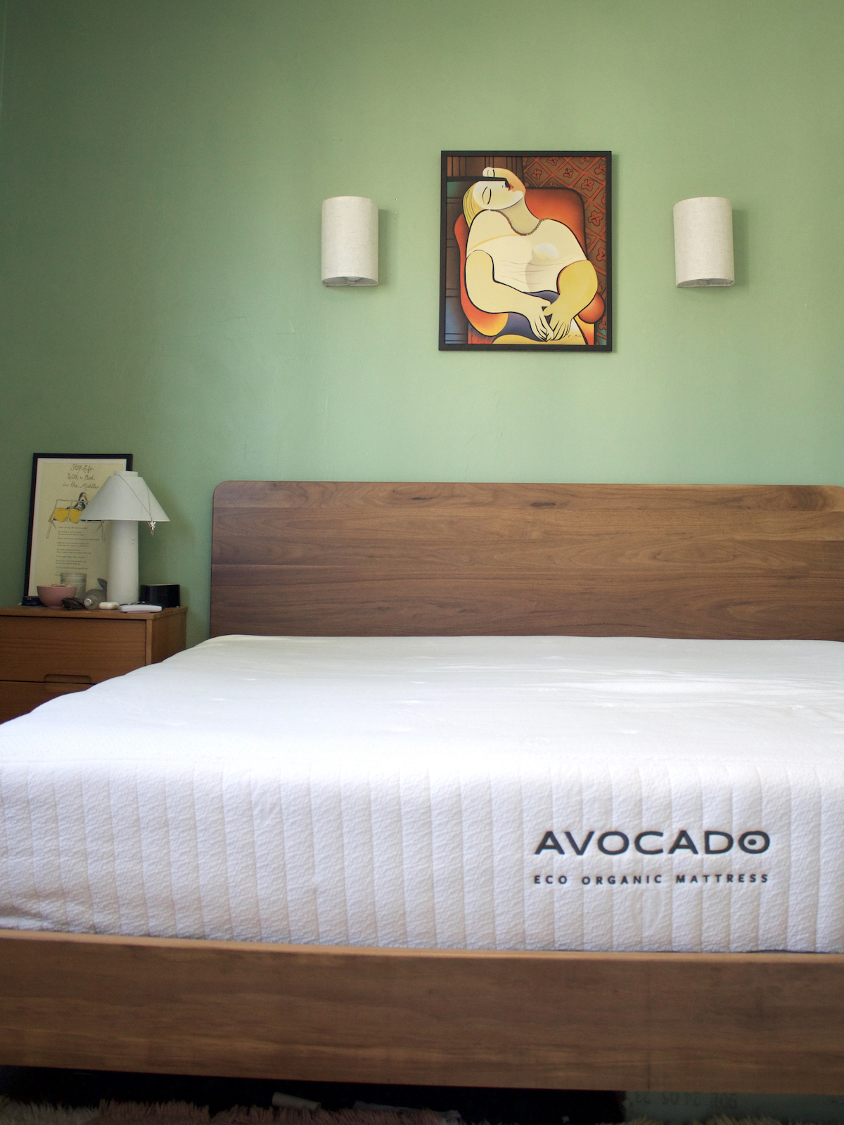 A neatly made bed with an Avocado latex mattress, wooden headboard, green wall, framed art, two wall sconces, and a bedside table with a lamp and framed print.