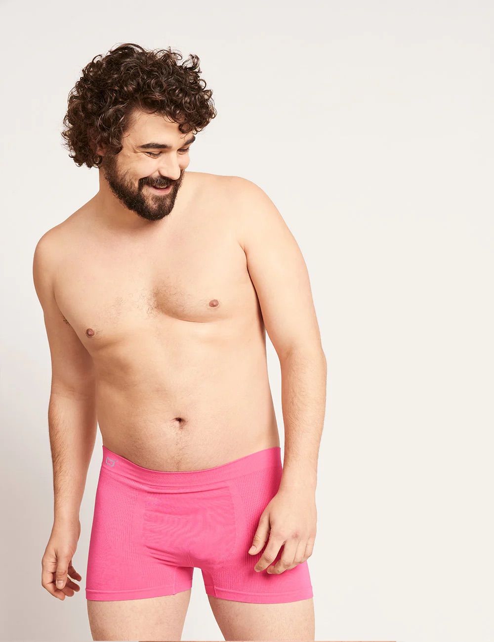 A man with curly hair and a beard stands against a plain background, wearing only bright pink boxer briefs and looking to his left.