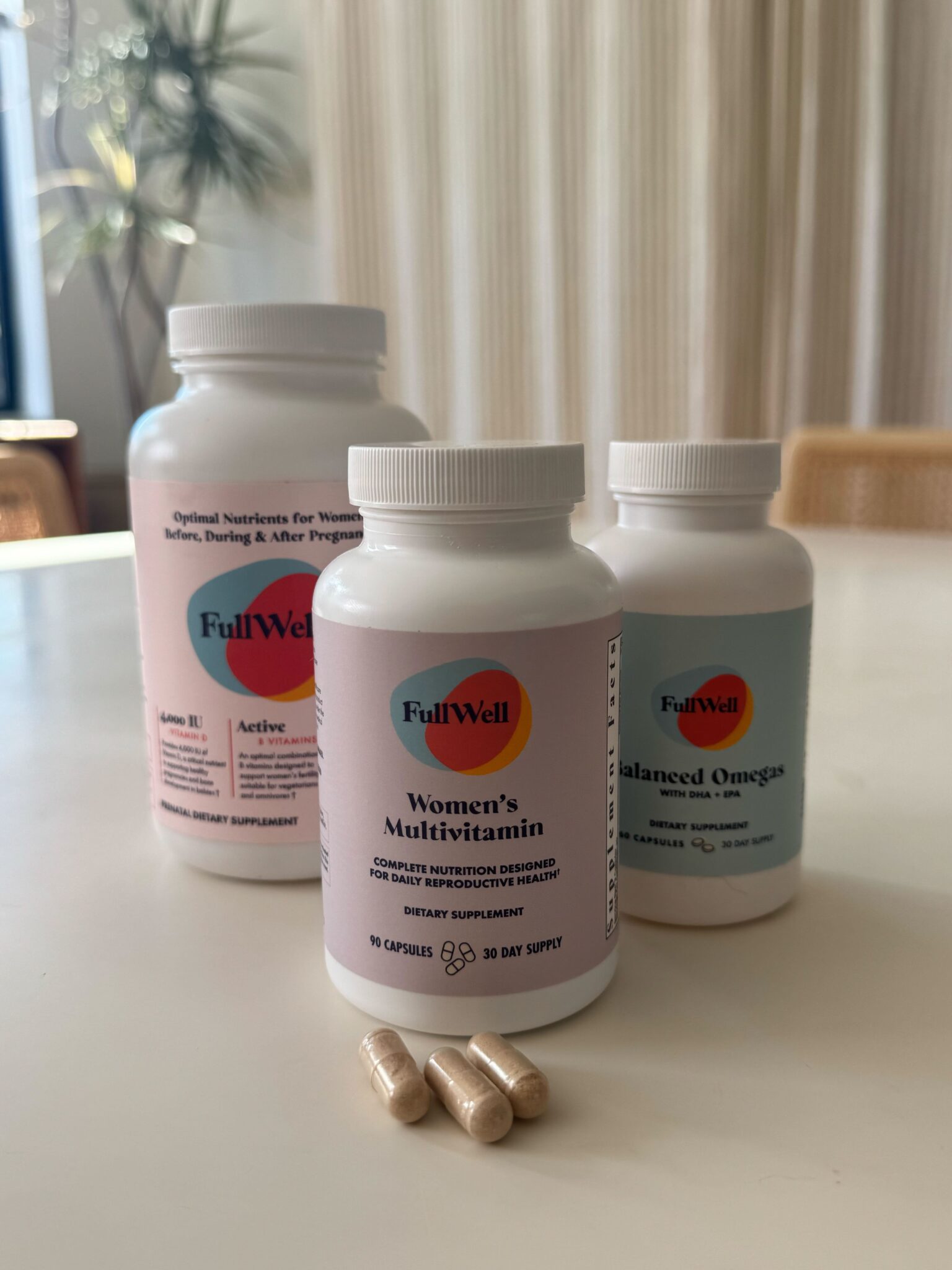 Three bottles of FullWell dietary supplements, including Women's Multivitamin, are on a table with three beige capsules in front of them.
