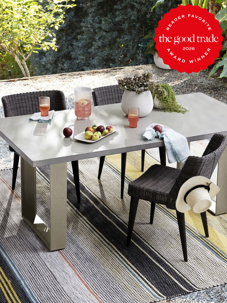 A modern outdoor dining table with four chairs, fruit, drinks, a pitcher, and a vase, set on a striped rug in a garden. A straw hat hangs on one chair.