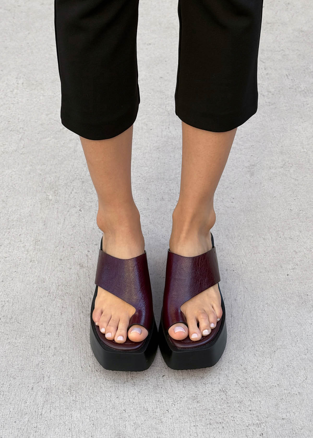 Person wearing black pants and burgundy platform sandals standing on a light gray concrete surface.