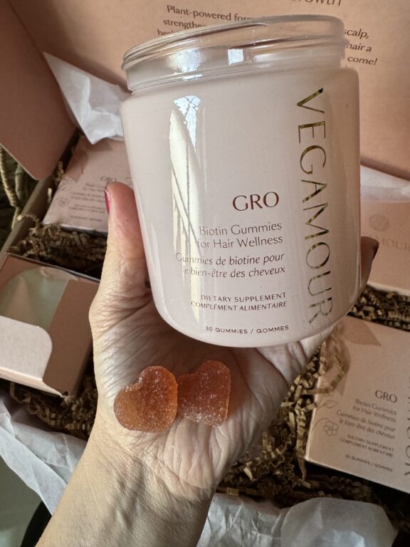 A hand holds a jar of Vegamour GRO Biotin Gummies for Hair Wellness with two heart-shaped gummies visible on the palm.