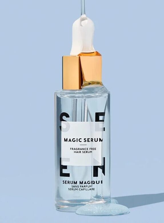 A clear bottle of Magic Serum hair serum with a dropper and gold cap, shown with spilled serum nearby. Text advises using 2-5 drops on mid-lengths to ends of hair.