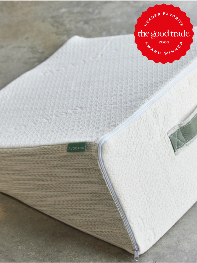 A wedge-shaped pillow with a fabric cover, zipper, and handle sits on a concrete floor. A red "The Good Trade 2026 Award Winner" badge is in the top right corner.