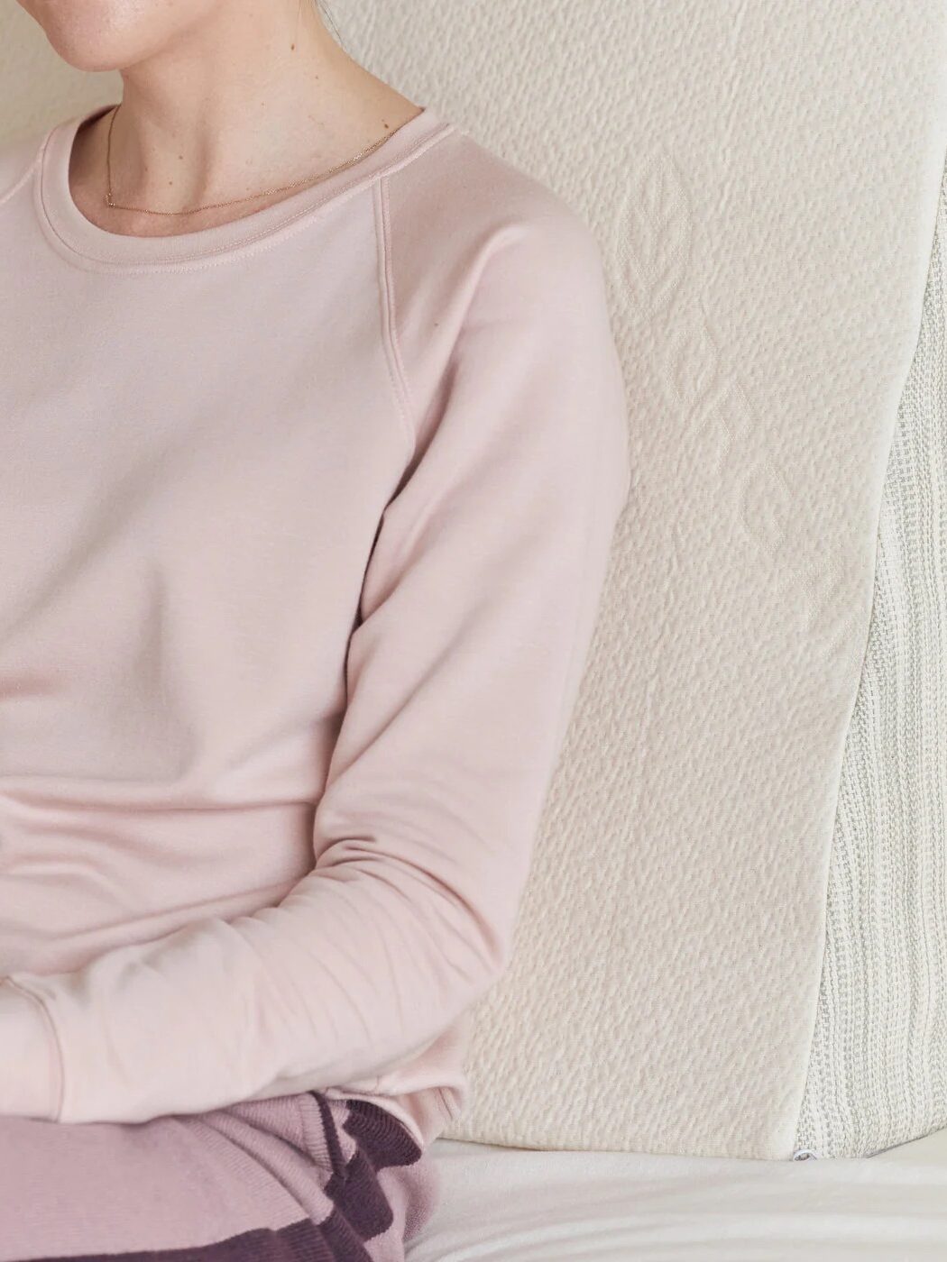 Person in a light pink long-sleeve shirt sits on a bed, holding a book, with a textured cream backrest cushion behind them.