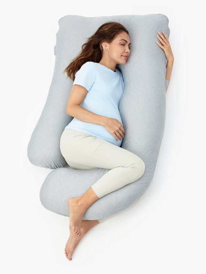 A woman lies on her side, hugging a large, U-shaped gray body pillow against a white background.