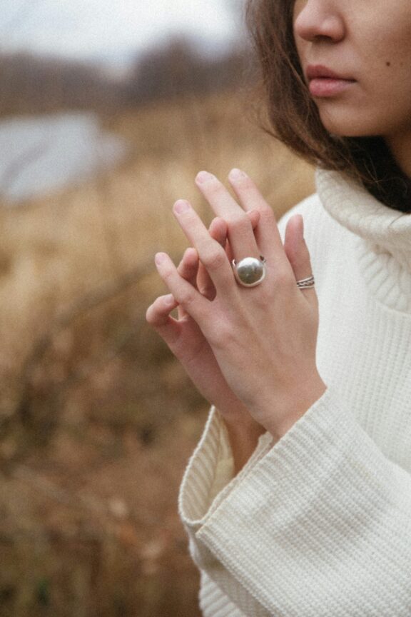 A person in a cream sweater outdoors touches their fingers, wearing a large silver ring and a thin band ring, with a blurred natural background.