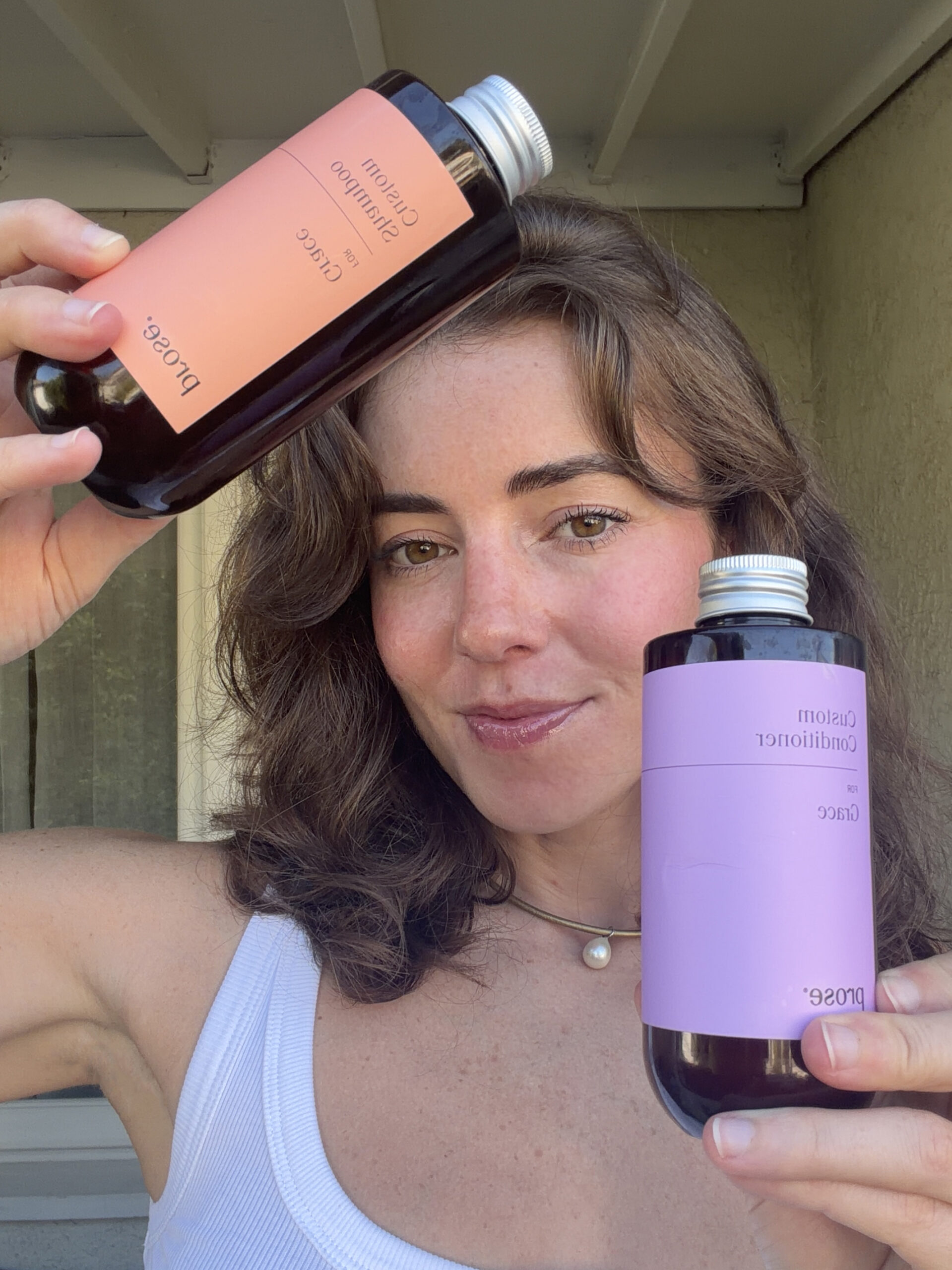 Woman in a white tank top holds two bottles labeled “Custom Shampoo” and “Custom Conditioner” by Prose while standing outside.