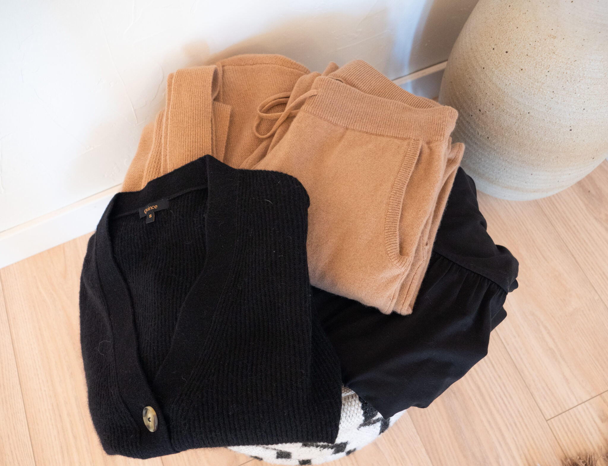 A folded black cardigan, black pants, and tan loungewear set sit on a woven basket on a light wood floor next to a beige ceramic vase.