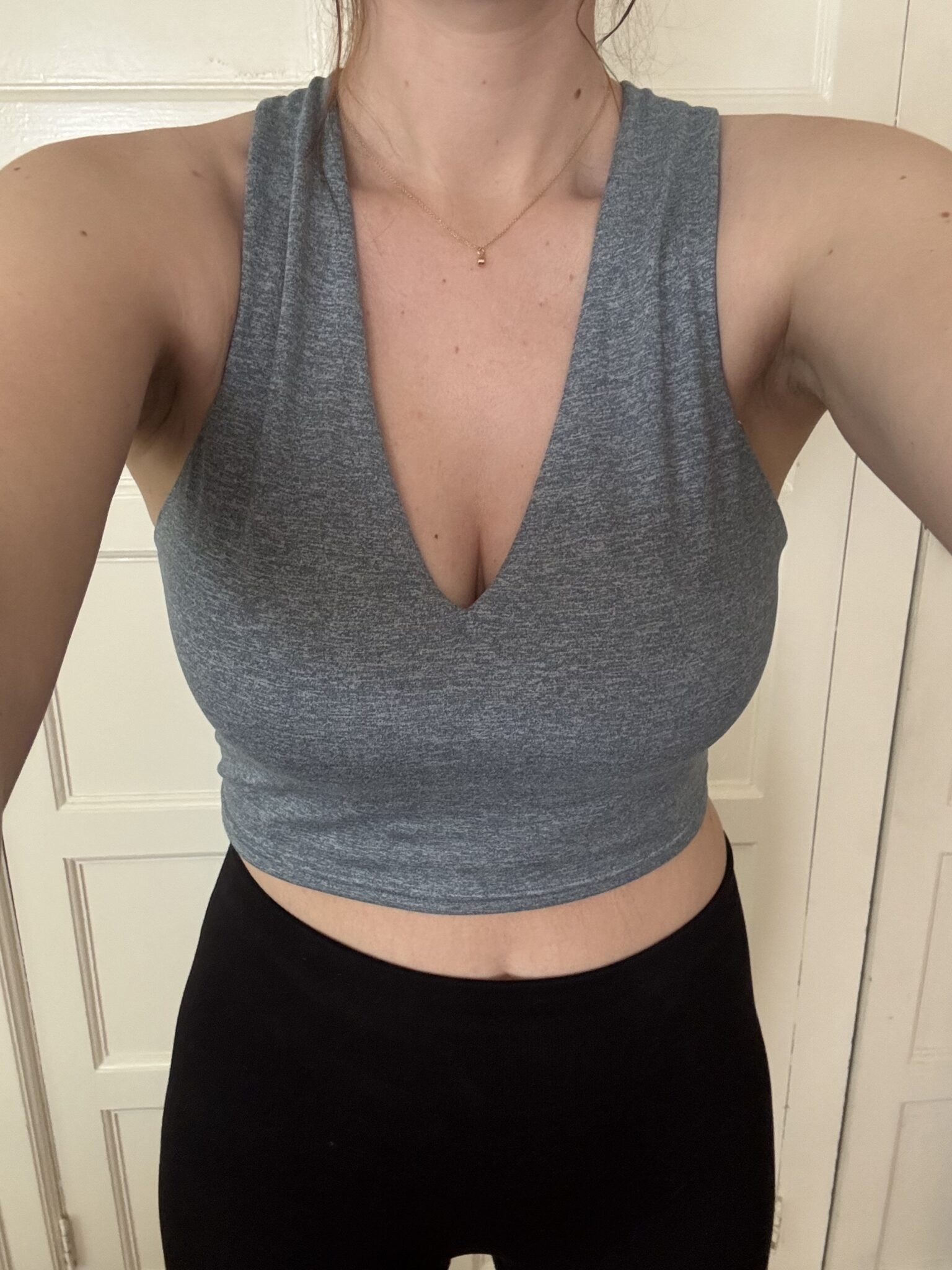 Person wearing a sleeveless gray crop top and black pants standing indoors in front of a cream-colored door.