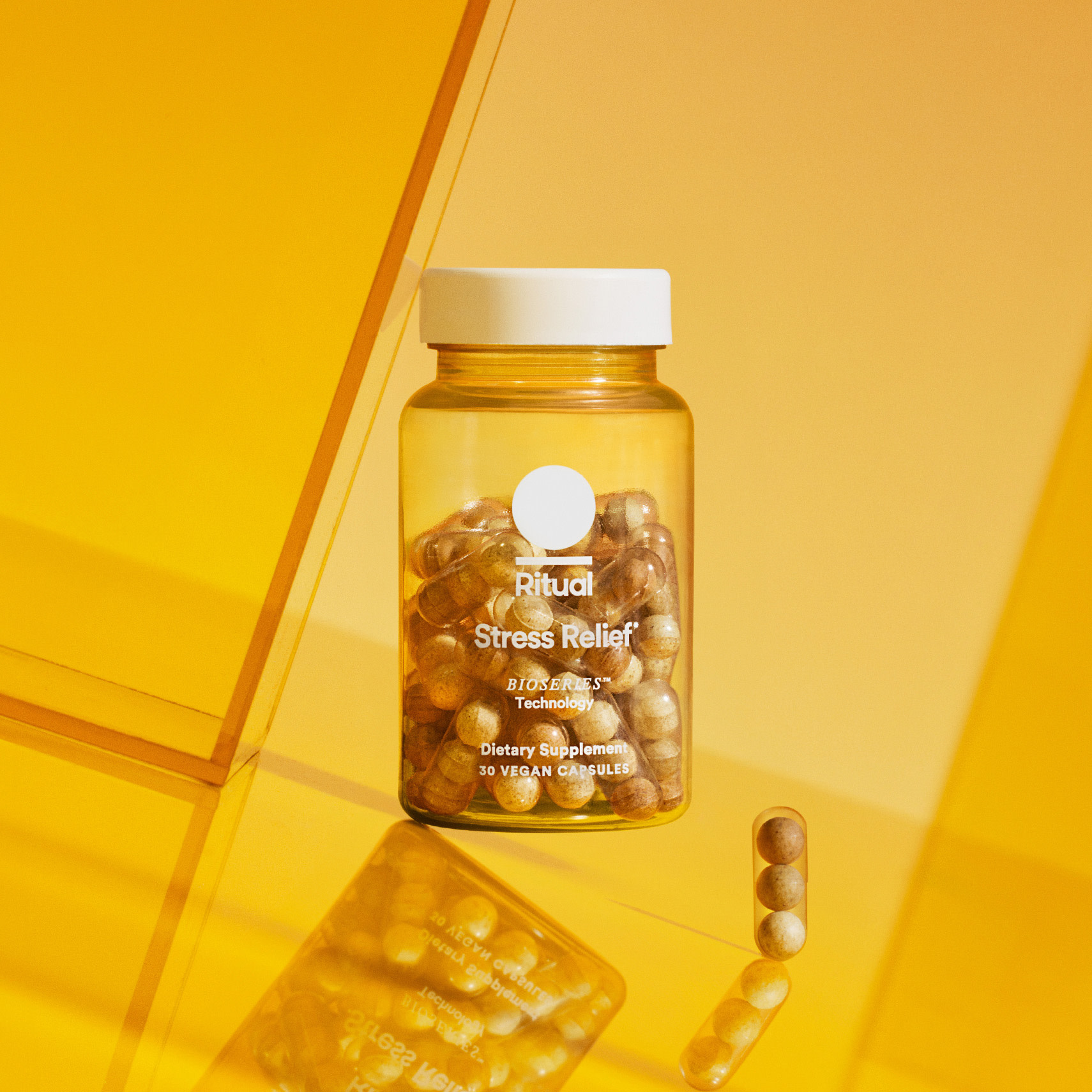 A clear bottle labeled "Ritual Stress Relief" containing capsules, with a few capsules placed outside, set against a yellow background.