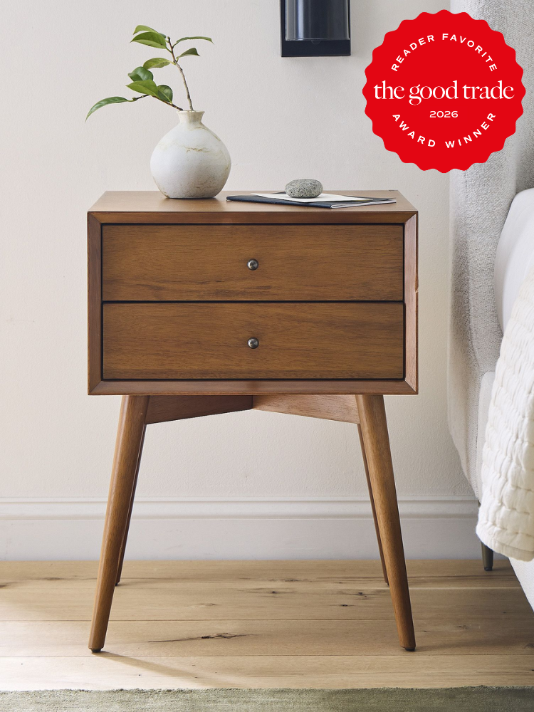 Mid-century modern wooden nightstand with two drawers, a ceramic vase with a plant, and a stone on top. "The Good Trade 2026 Award Winner" badge in the upper right corner.