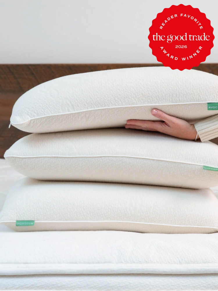 A person holding a stack of three white pillows on a bed with a red badge in the corner reading "the good trade award winner 2026.