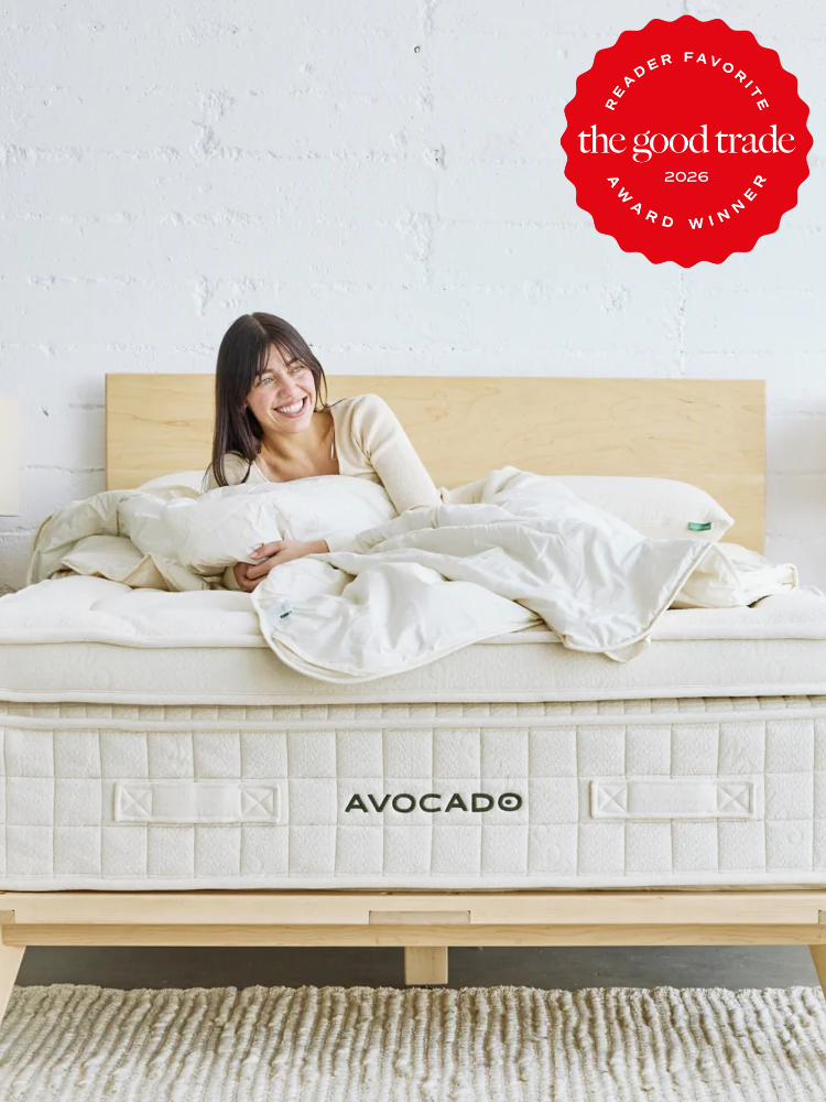 A woman sits smiling on an Avocado mattress in a bright bedroom. A red badge in the corner reads "The Good Trade Reader Favorite Award Winner 2026.