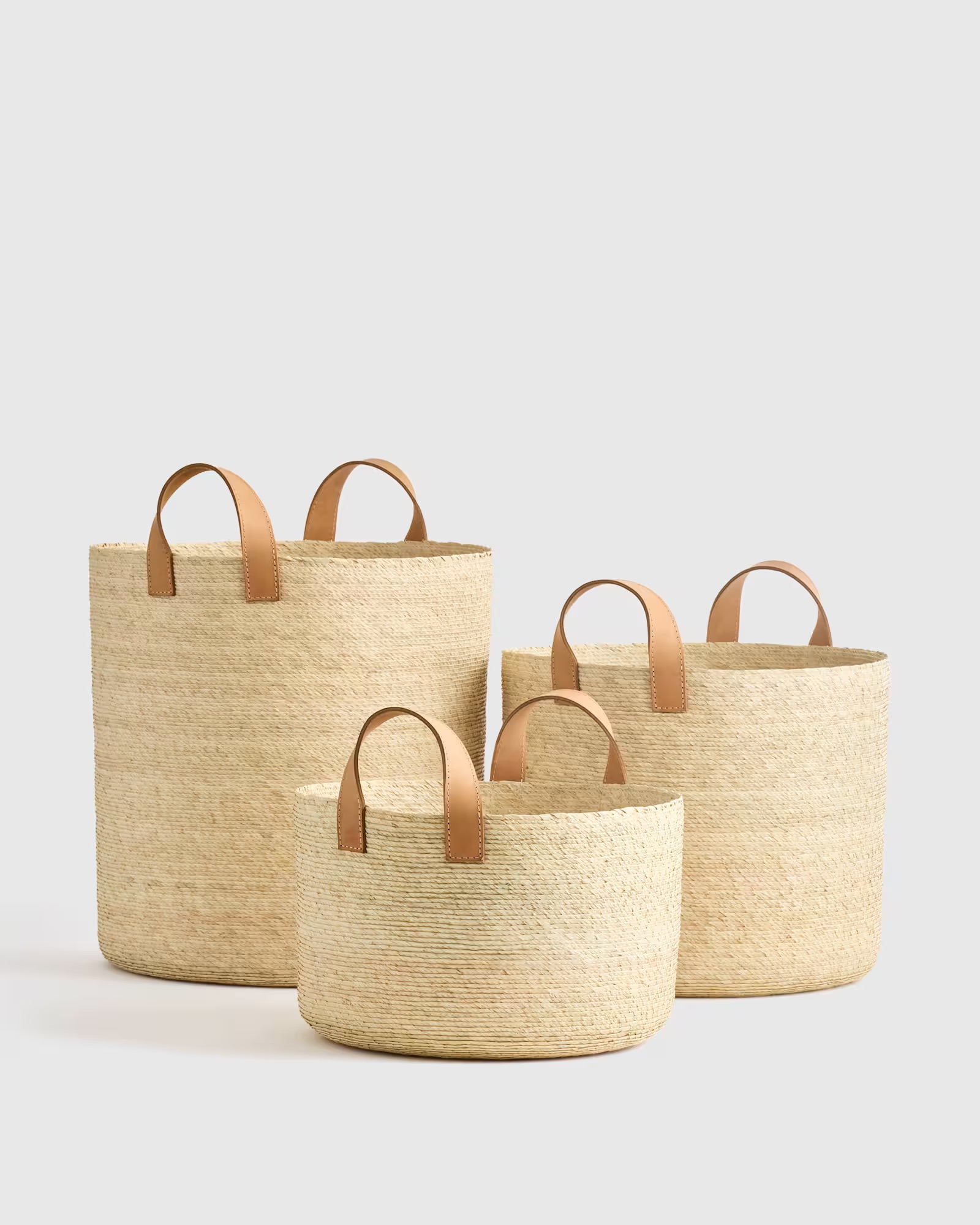 Three woven straw baskets with tan leather handles in small, medium, and large sizes, arranged on a light background.