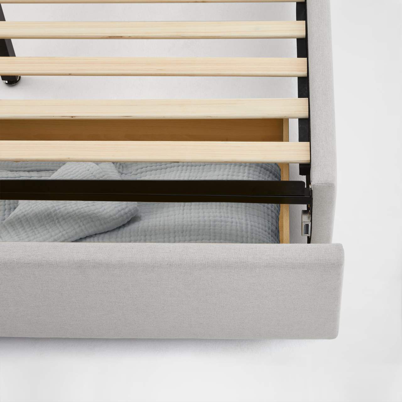 A close-up view of a bed frame with wooden slats, a fabric-upholstered headboard, and a gray textured blanket on the floor beneath.