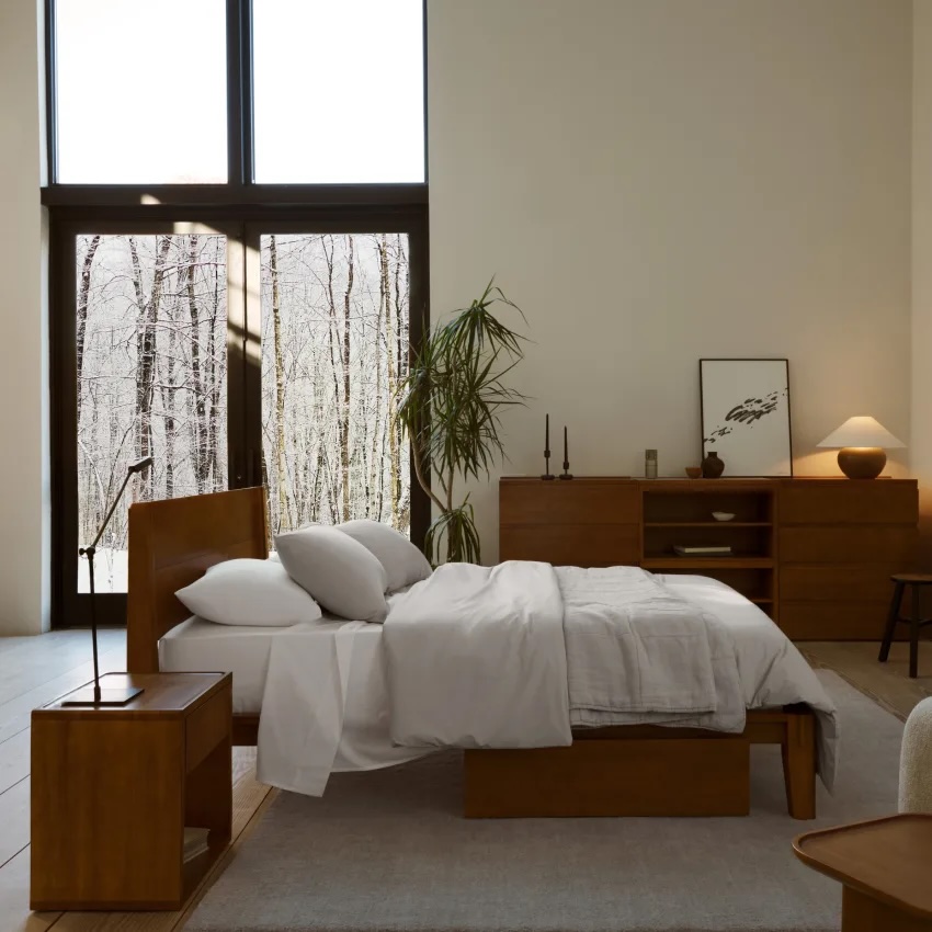 Minimalist bedroom with a wooden bed, white bedding, nightstand, dresser, potted plant, and large windows overlooking leafless trees.
