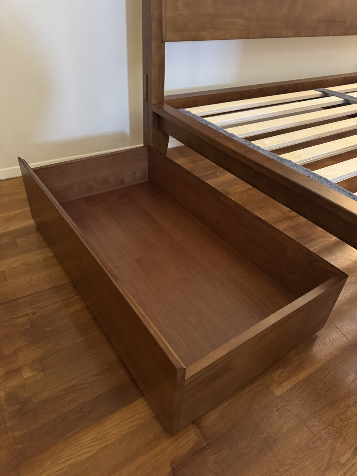 A wooden storage drawer is pulled out from under a wooden bed frame on a hardwood floor.