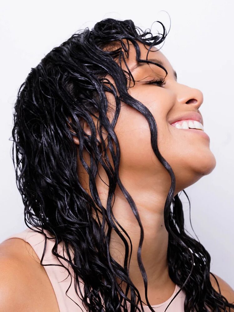 Woman with wet, curly black hair smiles with eyes closed, head tilted slightly back against a plain light background.