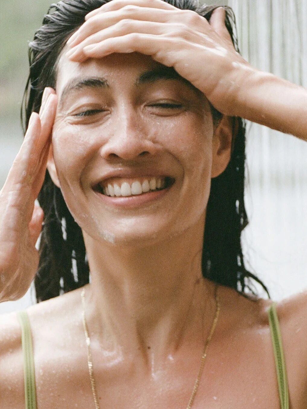 A person smiles while standing under running water outdoors, using their hands to wash their face.