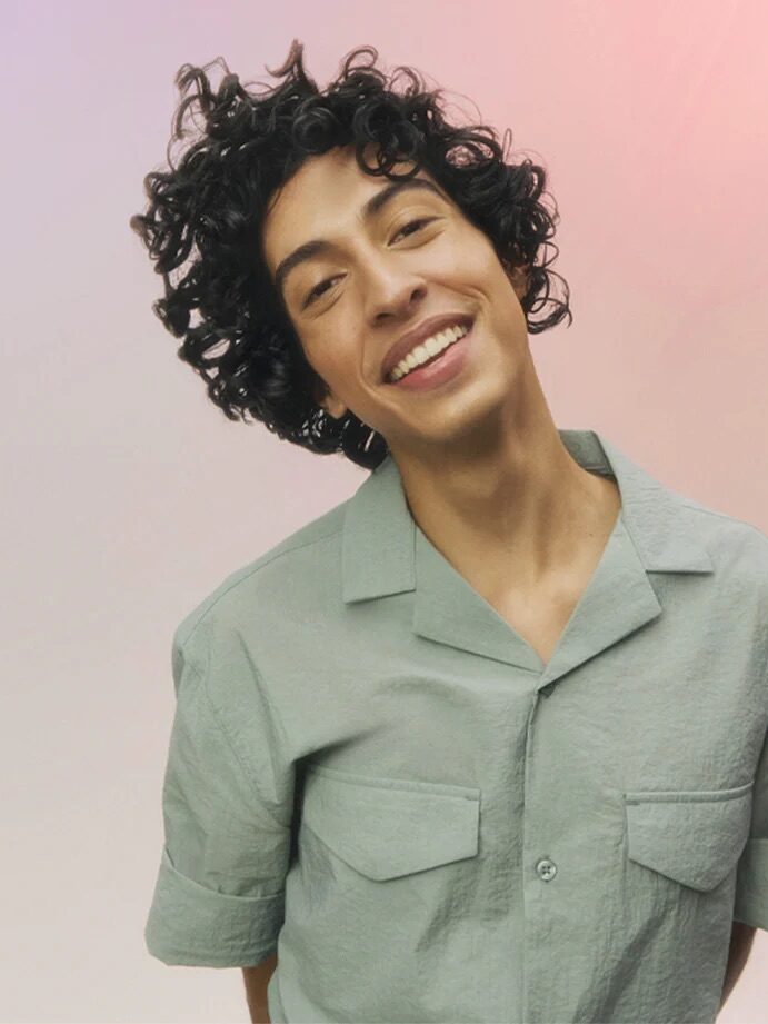 A person with curly hair, wearing a light green short-sleeved button-up shirt, is smiling against a pastel gradient background.