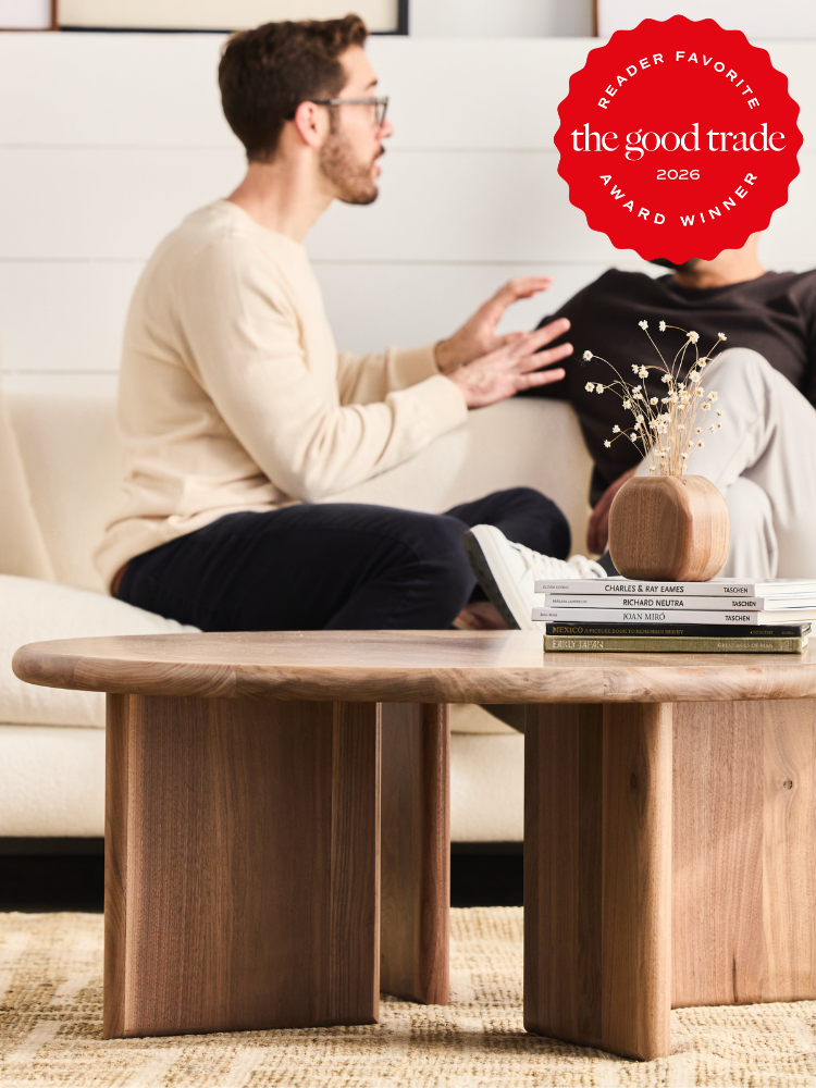 Two people sit on a couch having a conversation behind a wooden coffee table with books, a vase, and dried flowers. A red award badge is in the top right corner.