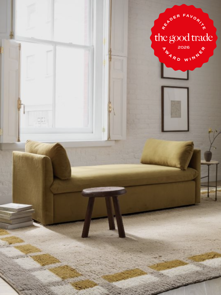 A minimalist living room with a mustard yellow daybed, small wooden stool, geometric rug, and a large window; an award badge is displayed in the corner.