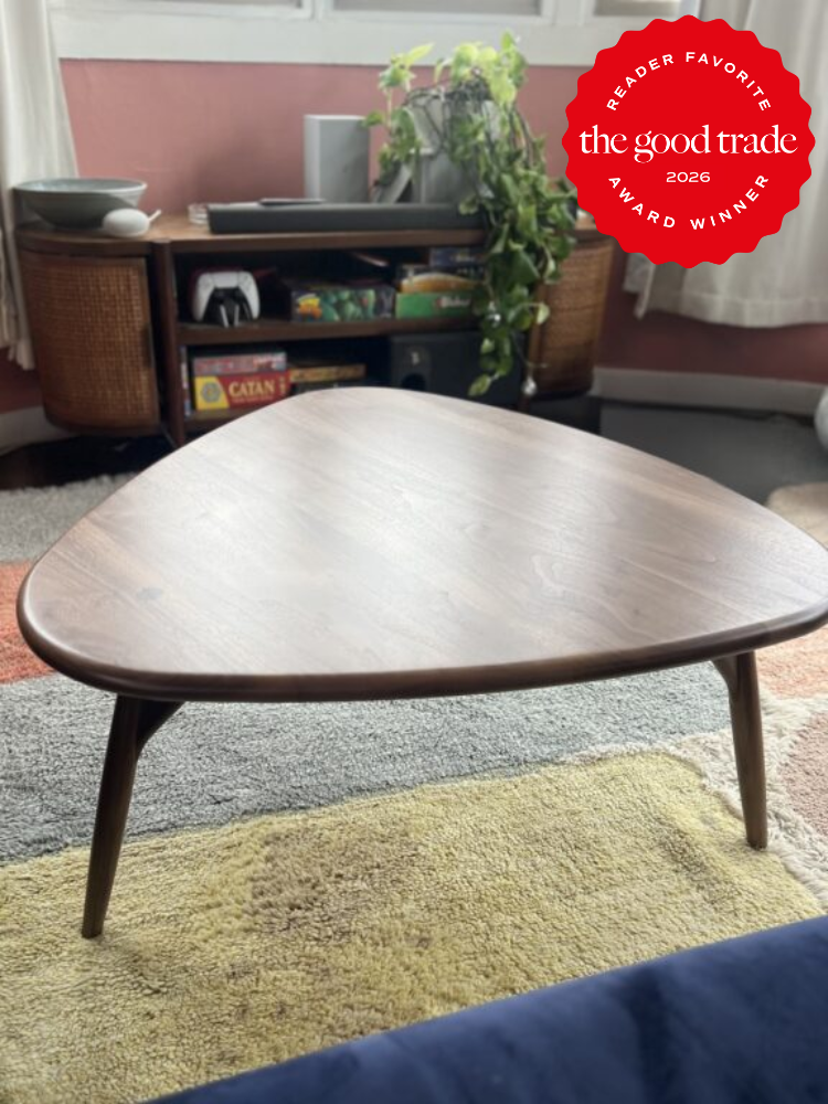 A triangular wooden coffee table on a multicolored rug in a living room, with shelves, a plant, and board games in the background. A red “The Good Trade” award badge is in the top right corner.