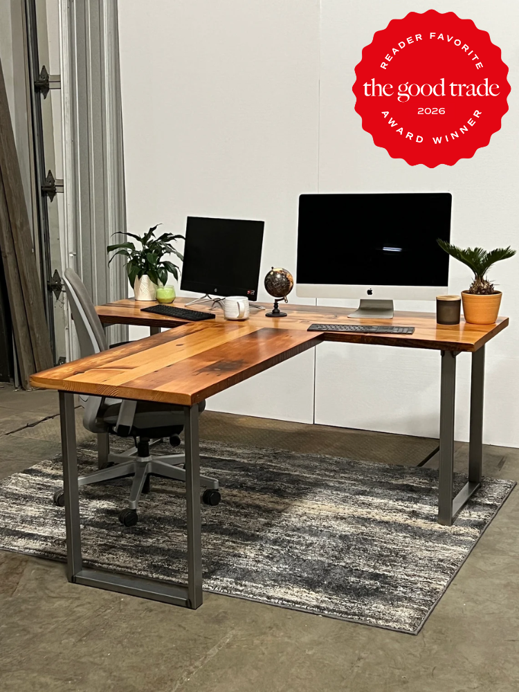 L-shaped wooden desk with two monitors, a chair, plants, and office supplies on a rug in an industrial-style room. "The Good Trade 2026 Award Winner" badge in top right corner.