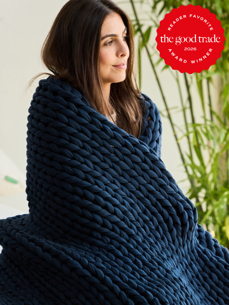 A woman sits wrapped in a chunky navy blue knit blanket. A "The Good Trade Award Winner 2026" badge appears in the top right corner.