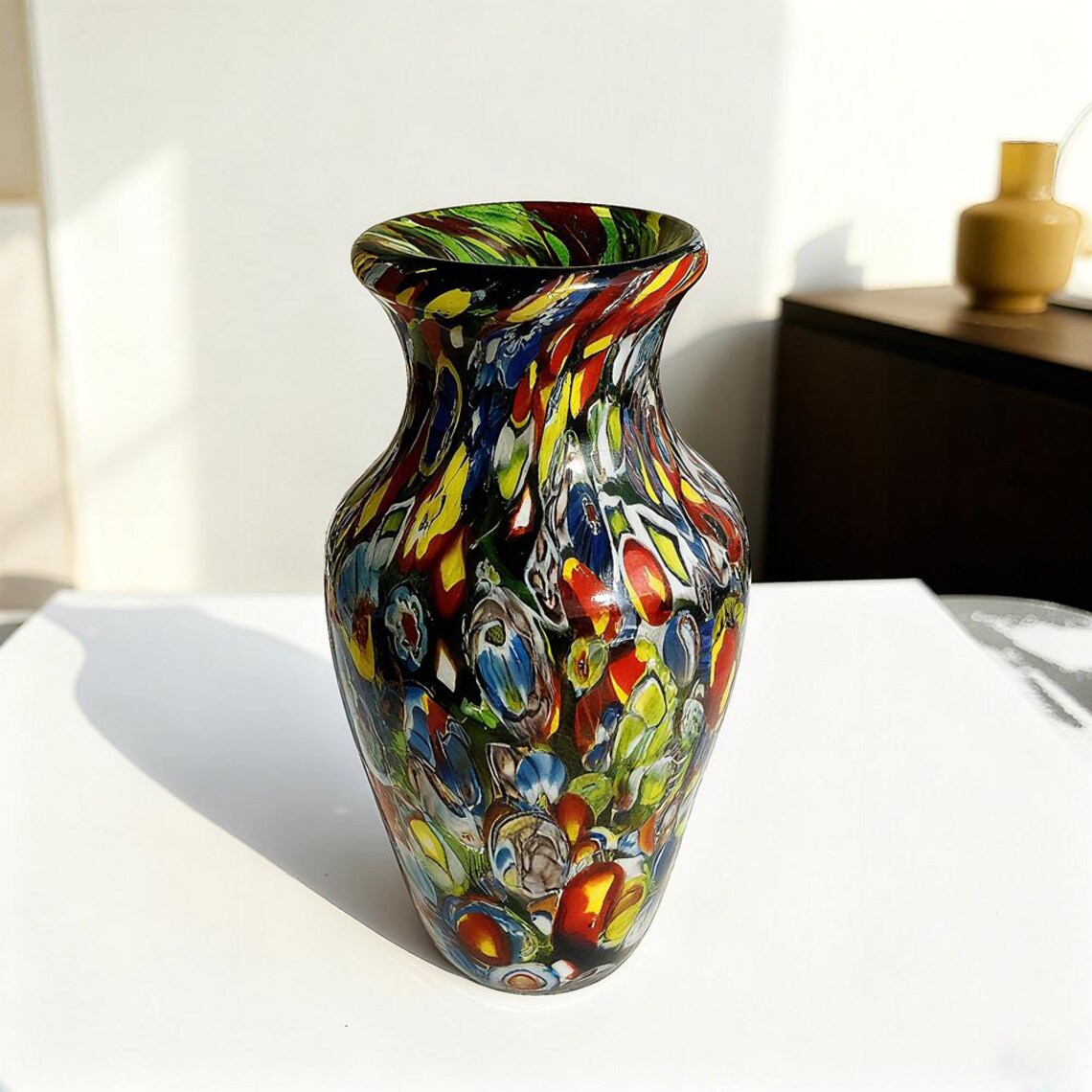 A colorful, patterned glass vase with a flared rim sits on a white surface in a sunlit room, with a yellow vase and dark furniture in the background.