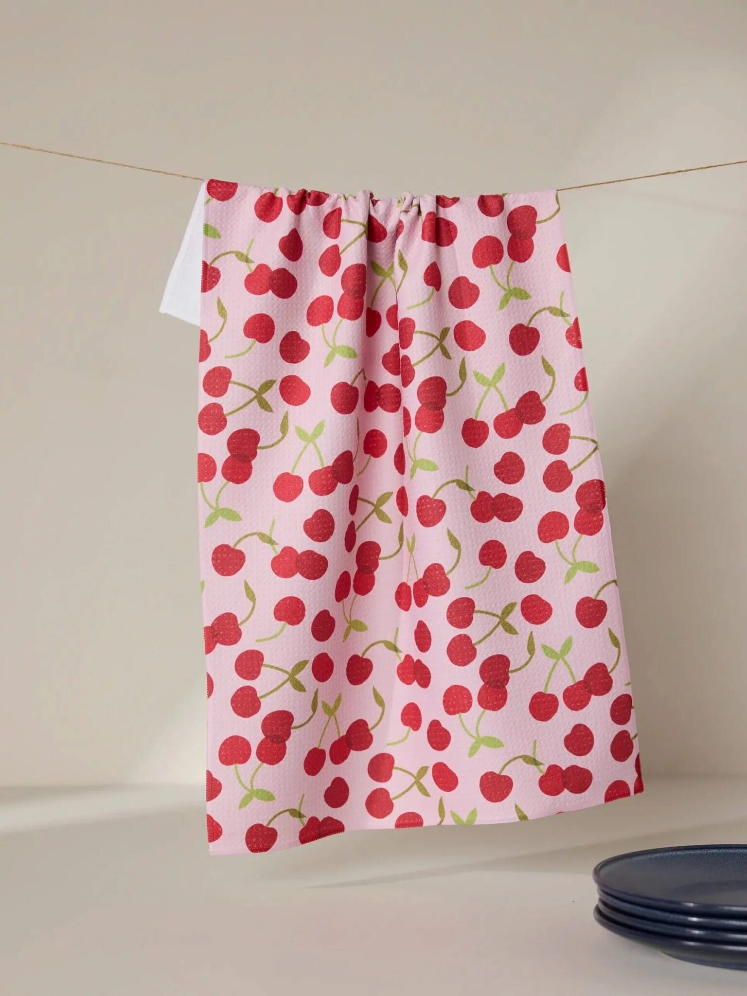 A white dish towel with a red cherry pattern hangs on a clothesline; part of a dark dish is visible in the lower right corner.