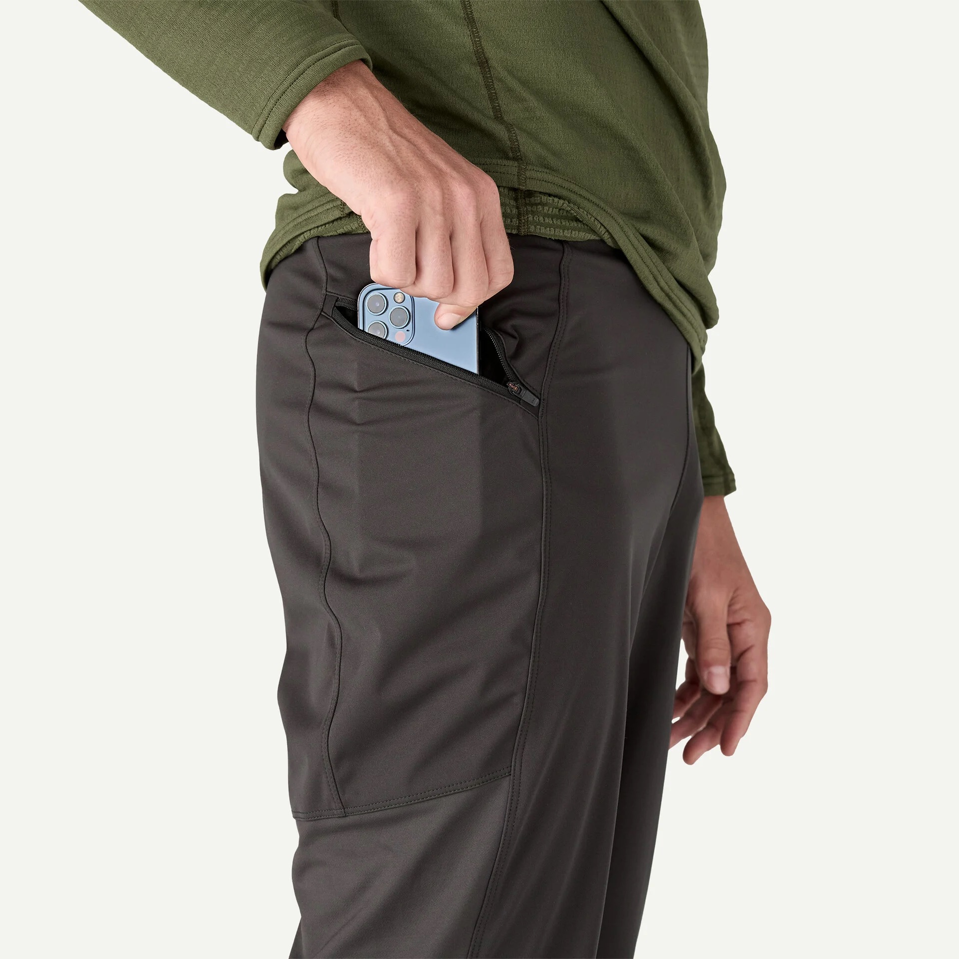 A person places a smartphone into the zippered side pocket of dark athletic pants while wearing a green long-sleeve shirt.