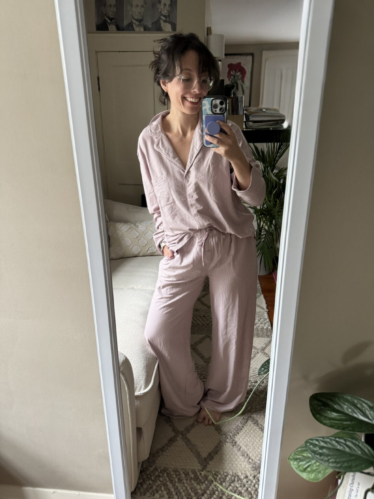 Person wearing light pink pajamas takes a mirror selfie in a living room with plants, a couch, and framed pictures visible in the background.