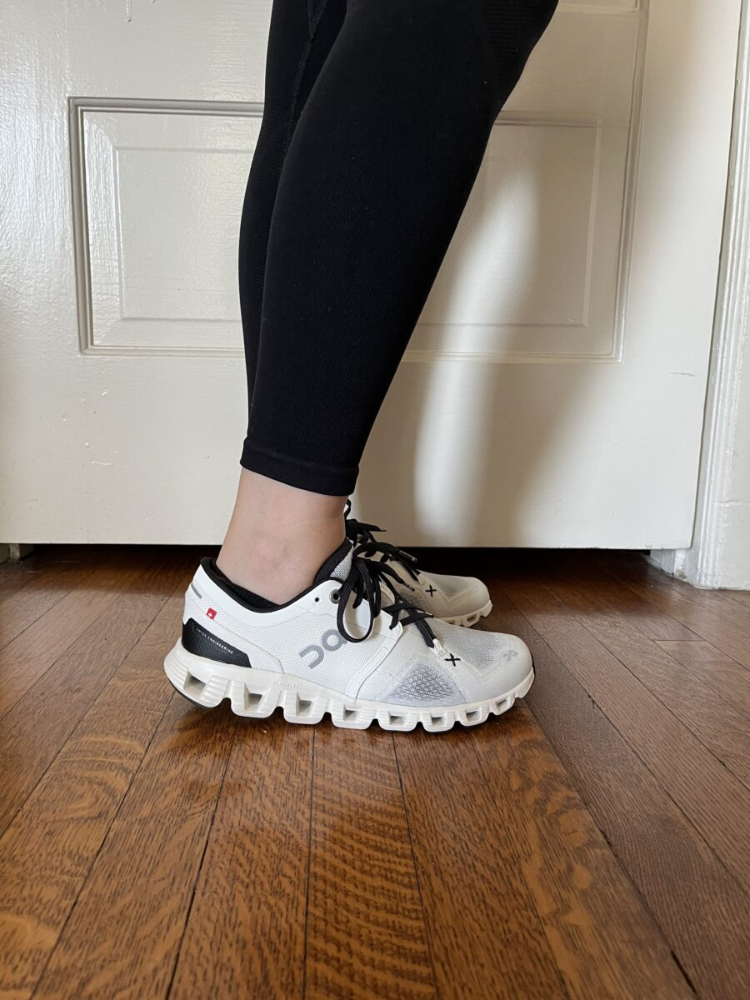 Person wearing black leggings and white On running shoes standing on a wooden floor in front of a white door.