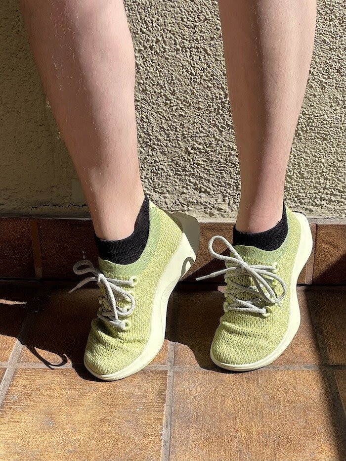 A person wearing light green knit sneakers with white laces and black socks stands on a tiled floor in sunlight.