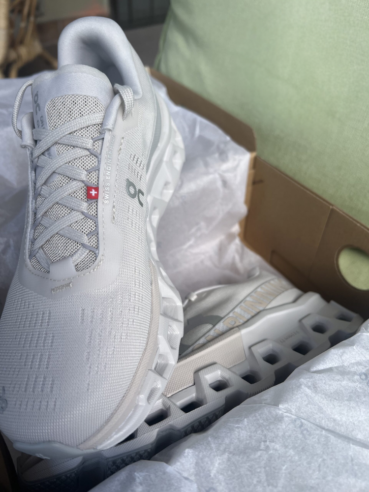 A pair of new white On running shoes sits in an open cardboard box lined with white tissue paper.