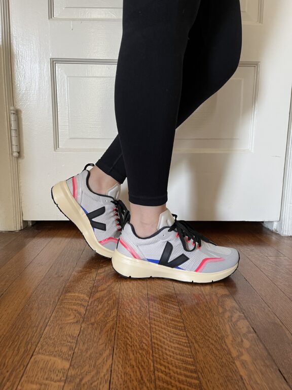 Person wearing black leggings and gray, red, and blue athletic shoes standing on a wooden floor in front of a white door.