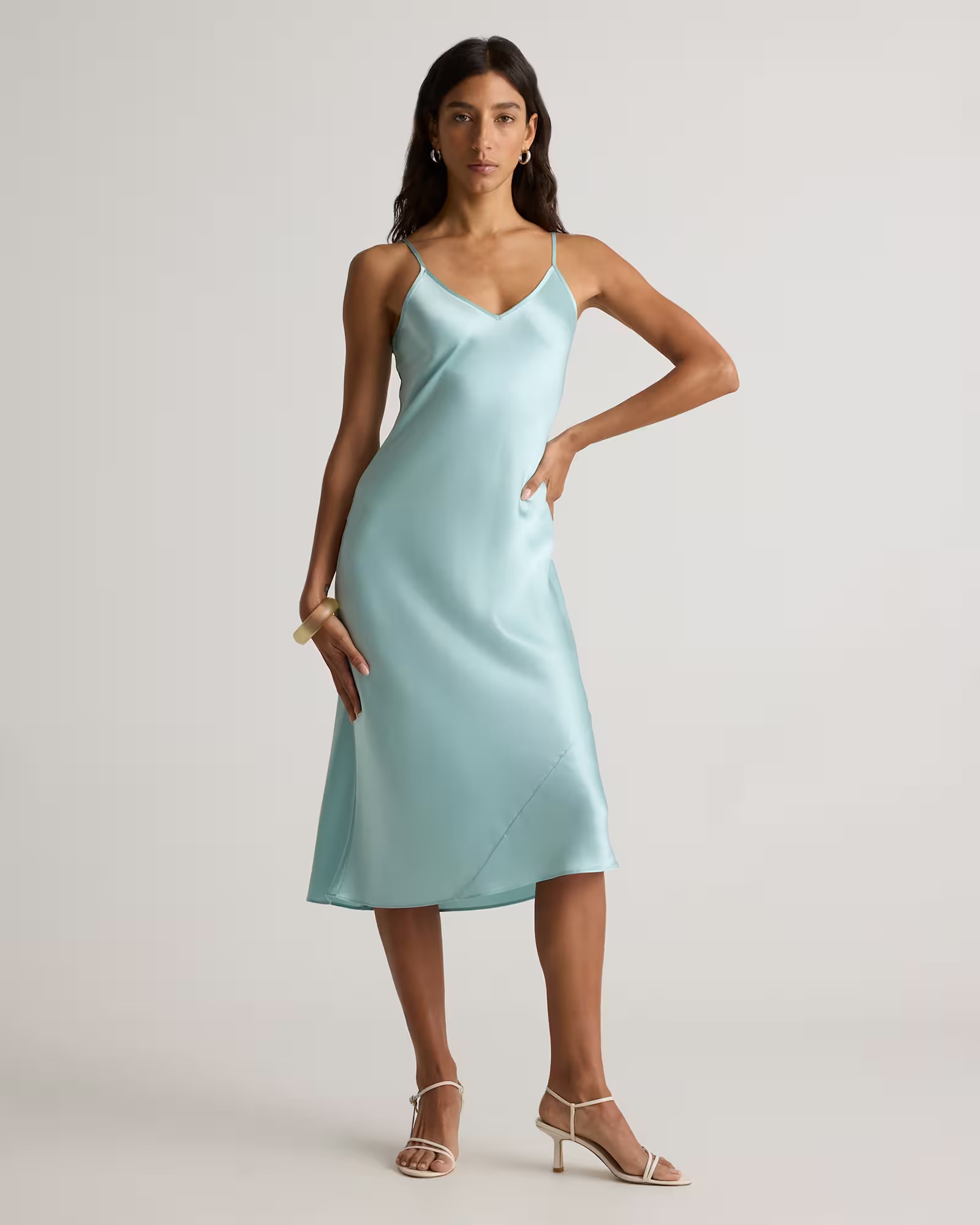 Woman wearing a light blue, sleeveless, midi-length slip dress with thin straps and beige heeled sandals, standing against a neutral background.
