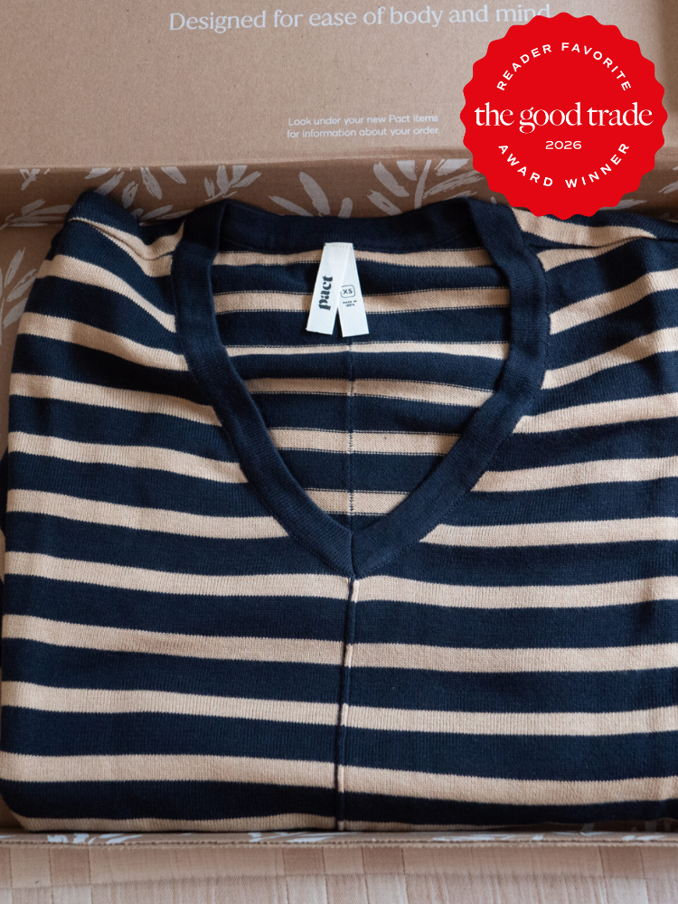 A folded navy and beige striped sweater with a V-neck and a white tag is placed inside an open cardboard box. A red “The Good Trade 2026 Award Winner” badge is on the top right.