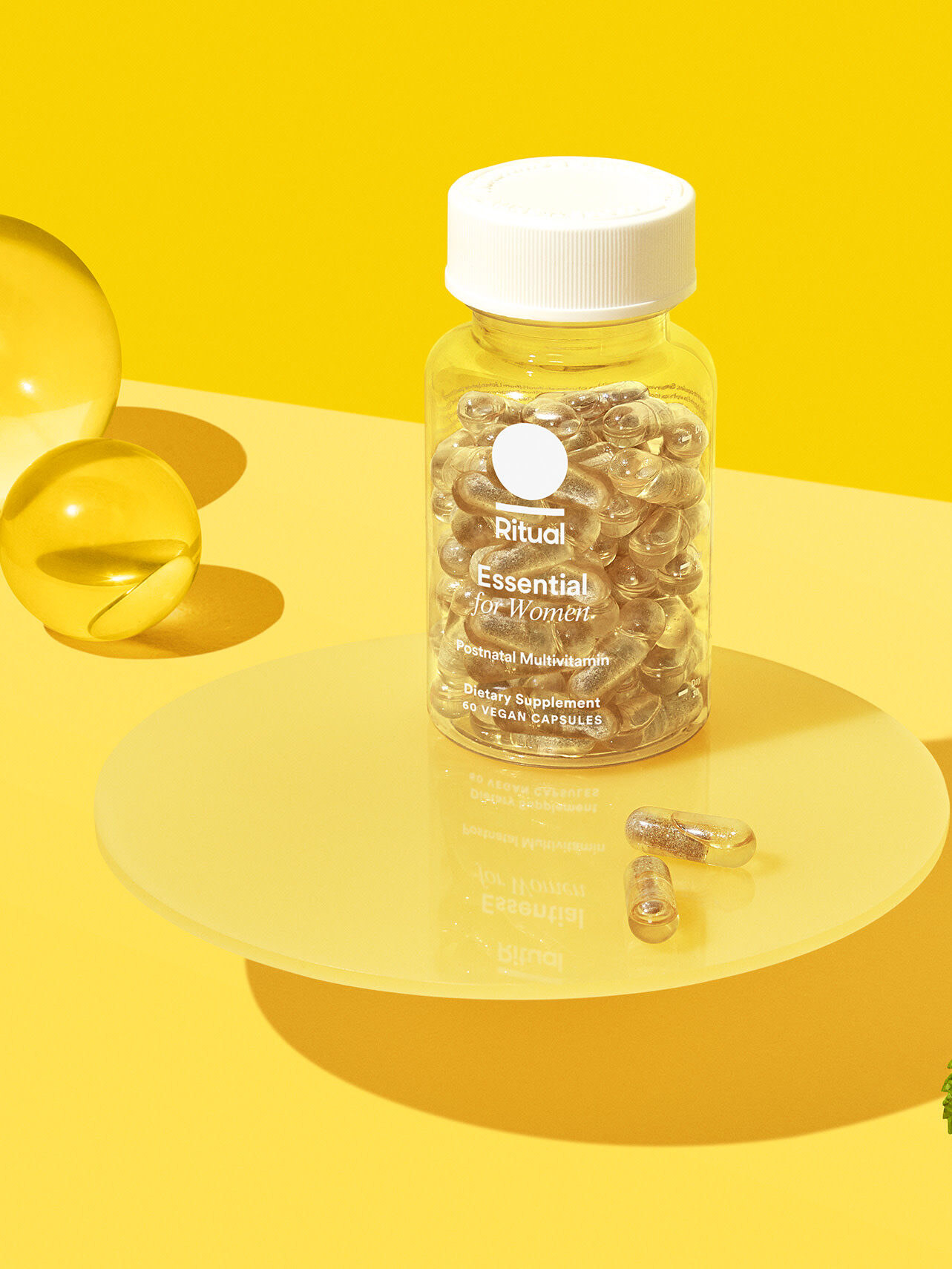 A clear bottle labeled "Ritual Essential for Women" multivitamin capsules is displayed on a round yellow surface with two loose capsules and a mint leaf nearby, against a yellow background.