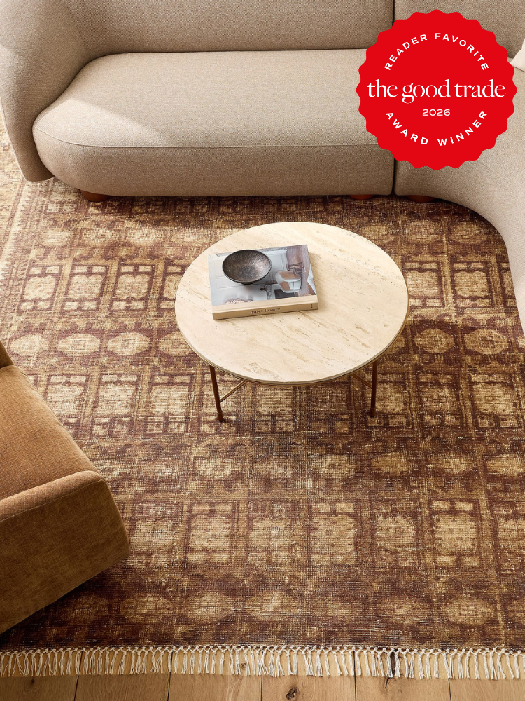 A round wooden coffee table with a book and ceramic plate sits on a patterned rug between a beige sofa and a brown chair; an award badge appears in the top right corner.